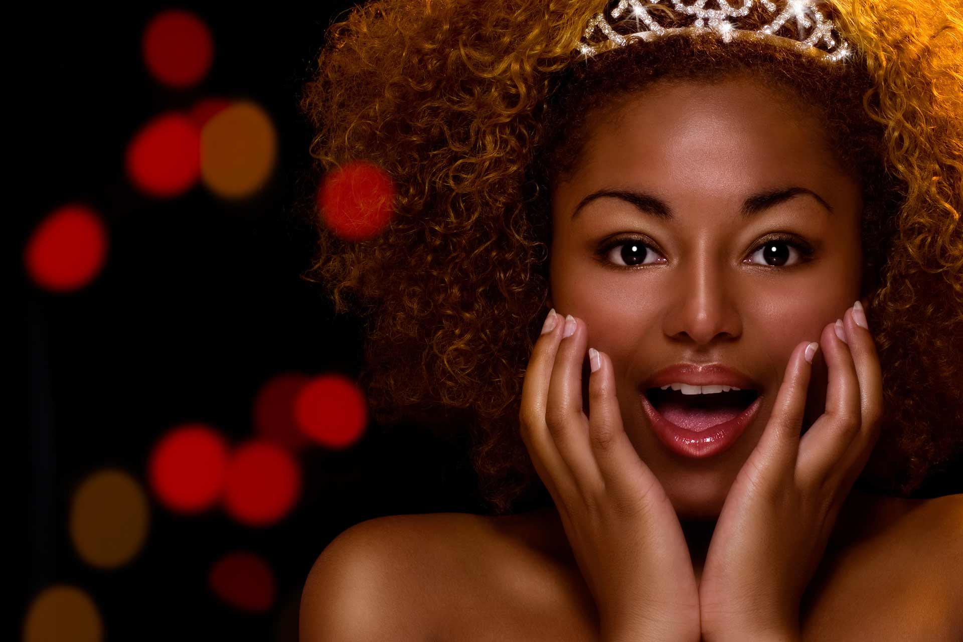 Black princess with hands on face in a surprise look; black background with red and gold blurred lights.