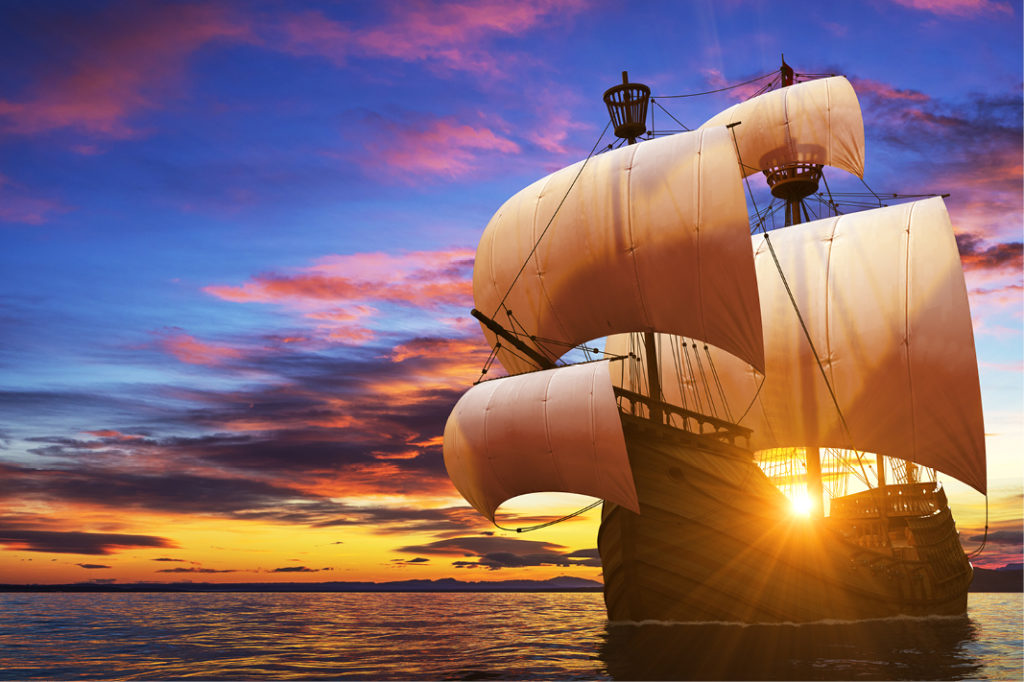 A wooden ship with four white sails on a blue ocean at sunset with pink clouds
