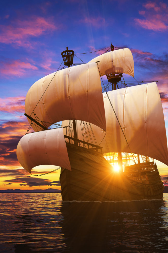 A wooden ship with four white sails on a blue ocean at sunset with pink clouds